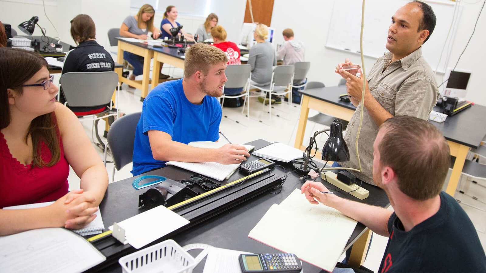 Students listening to professor as he visits their station in a physics lab.